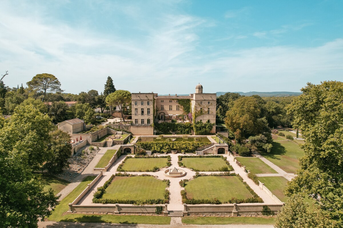 The exterior of a historic château is depicted, set among lush green trees and manicured gardens. The symmetrical garden layout includes pathways and a central fountain, providing a serene backdrop to the elegant building, which features multiple windows and a mix of architectural details.