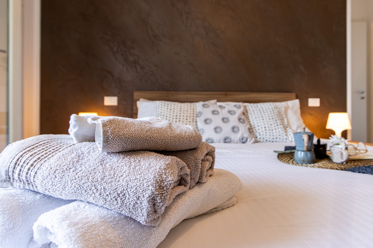 A neatly arranged set of towels sits atop a bed, complemented by patterned pillows against a textured wall. Soft lighting from bedside lamps creates a calm ambiance, while a small tray with cups and snacks is positioned on a woven placemat beside the bed.