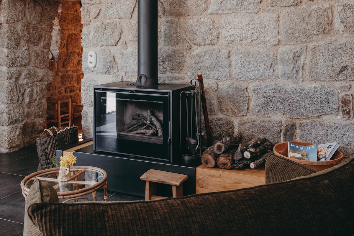 A cozy living area features a modern wood-burning stove, set against a backdrop of stone walls. A collection of firewood is neatly arranged nearby. A comfortable sofa and a small wooden side table enhance the inviting space, while a bowl of magazines adds a touch of leisure.