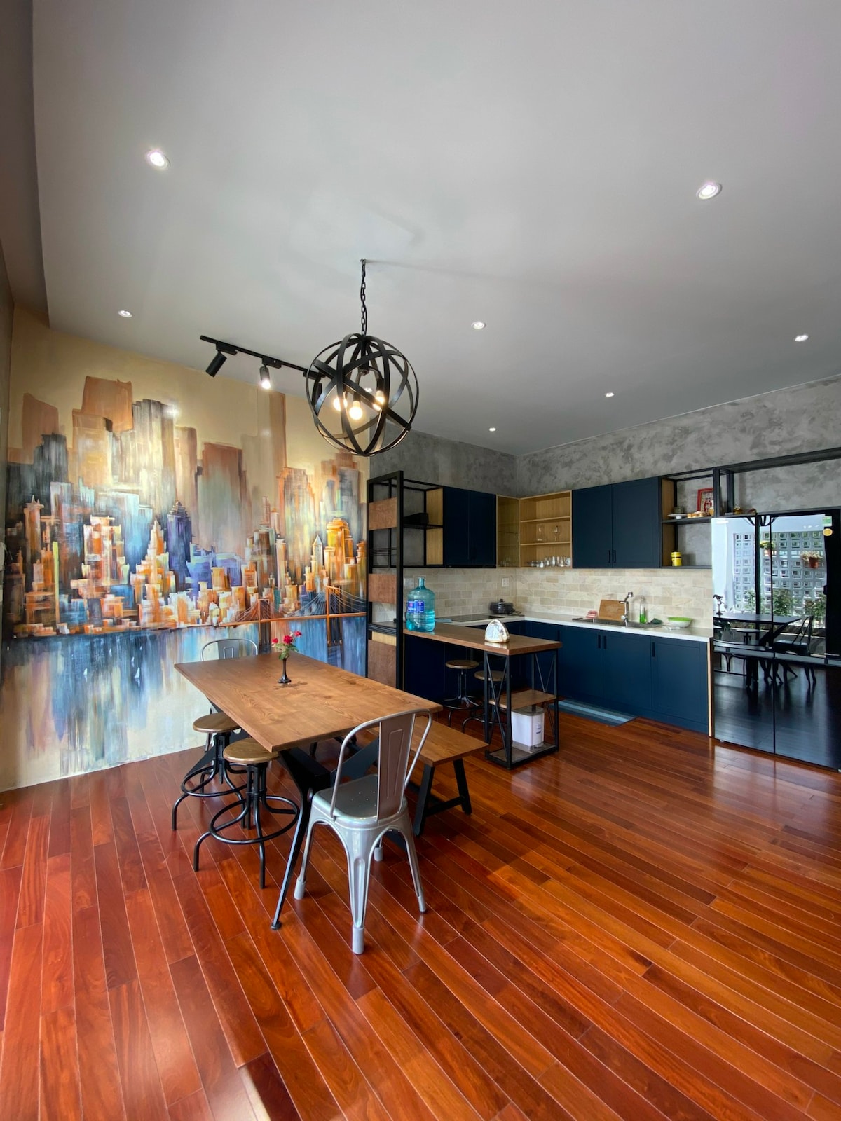 An open dining area features a wooden table surrounded by metal chairs. A striking cityscape mural adorns one wall, while modern cabinetry provides storage. Recessed lighting complements the warm tones of the hardwood floor, and a stylish light fixture hangs from the ceiling.