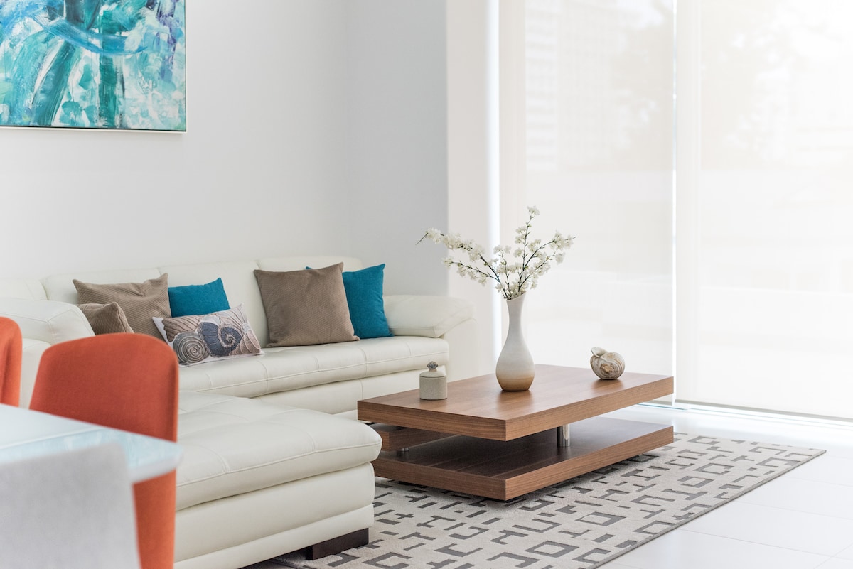 A modern living area features a light-colored sectional sofa adorned with pillows in blue and earth tones. A wooden coffee table sits at the center, accompanied by a decorative vase with flowers and a small sculpture. Natural light fills the space through large windows.
