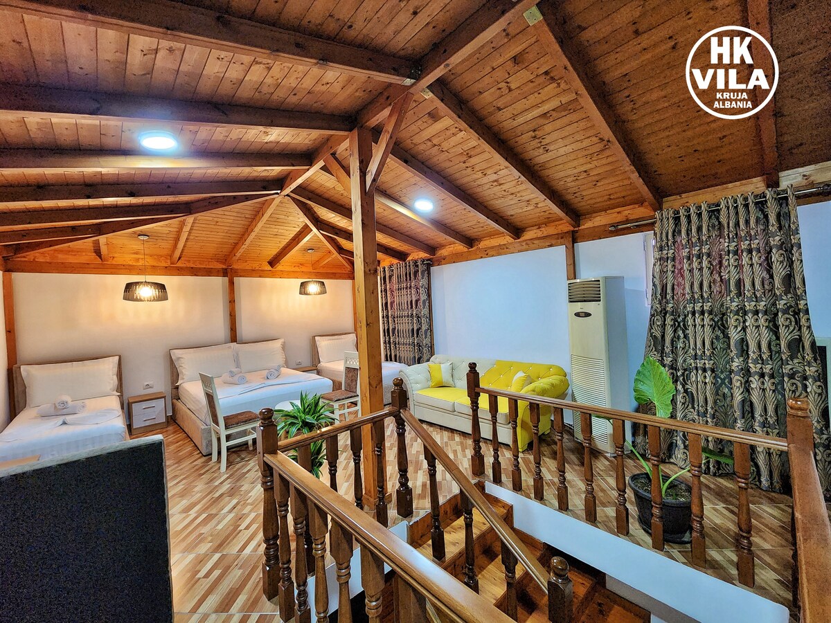 A spacious family suite features wooden beams and a staircase leading to an upper level. Multiple sleeping arrangements include two beds and a yellow couch. Decorative plants are positioned around the room, with windows dressed in patterned curtains allowing natural light to enter.