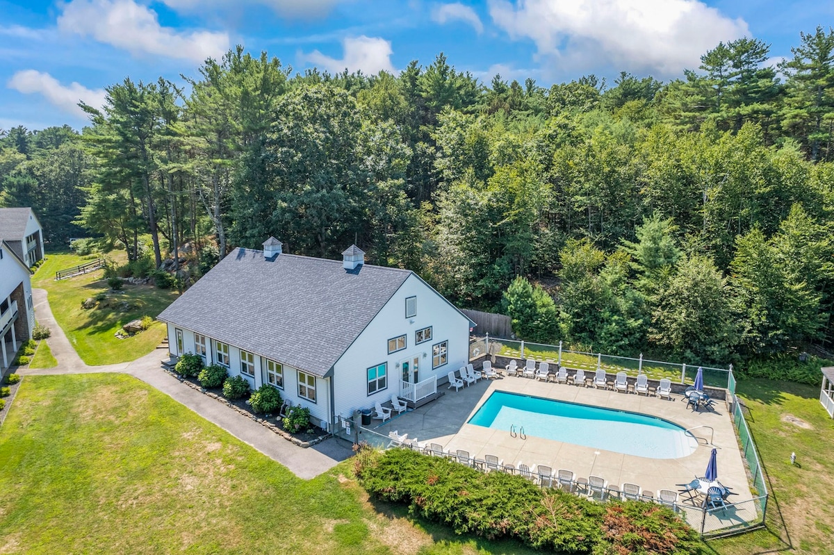 An aerial view of the resort showcases the outdoor pool surrounded by a spacious patio with lounge chairs. Lush greenery frames the property, while the building features multiple windows and an accessible layout. The area is designed for relaxation and recreation.