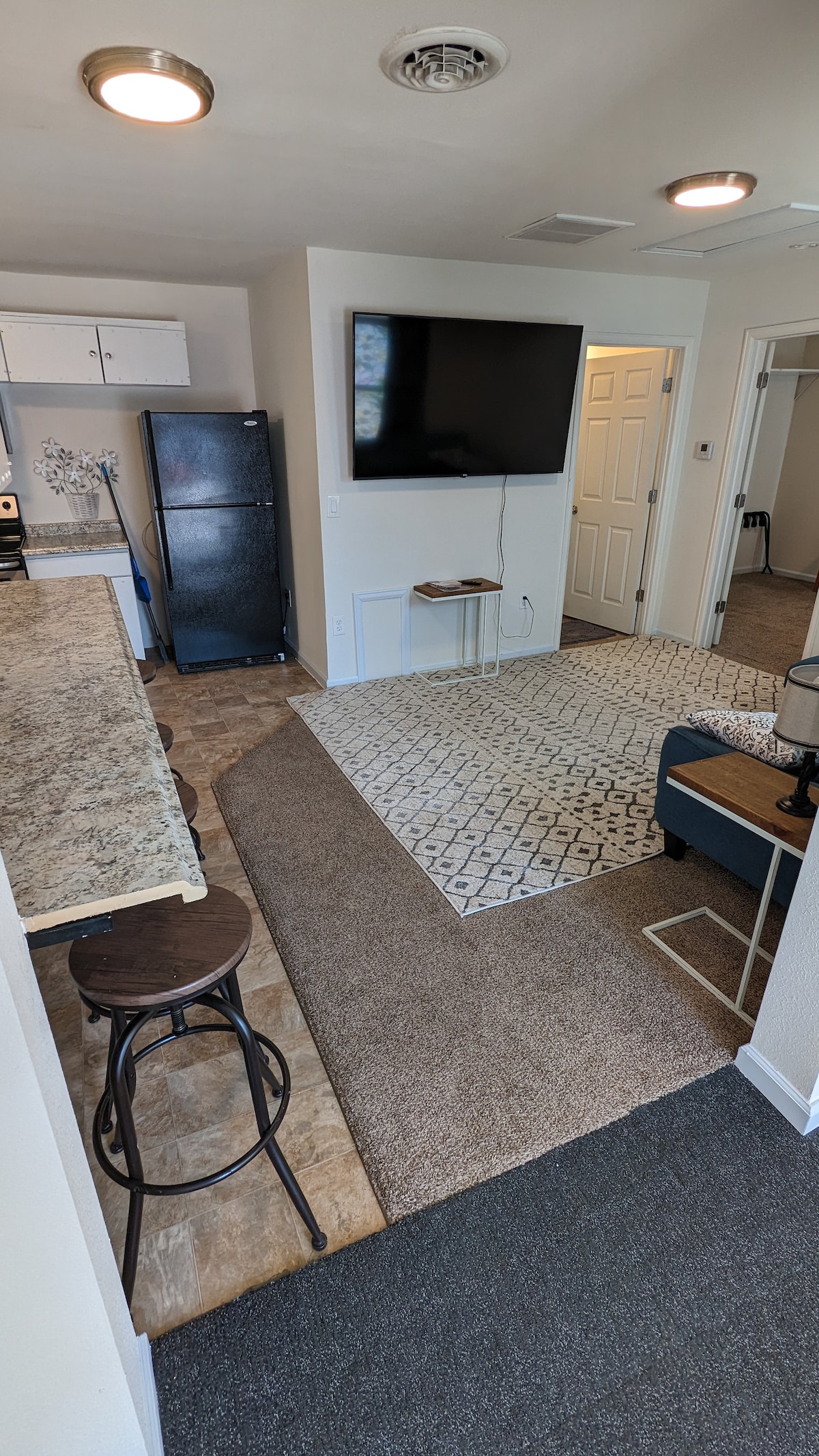 A cozy living area showcases a large flat-screen TV mounted on the wall. An inviting area rug sits under a low coffee table, while a bar counter with stools separates the living space from the kitchen. Natural light filters through nearby windows.