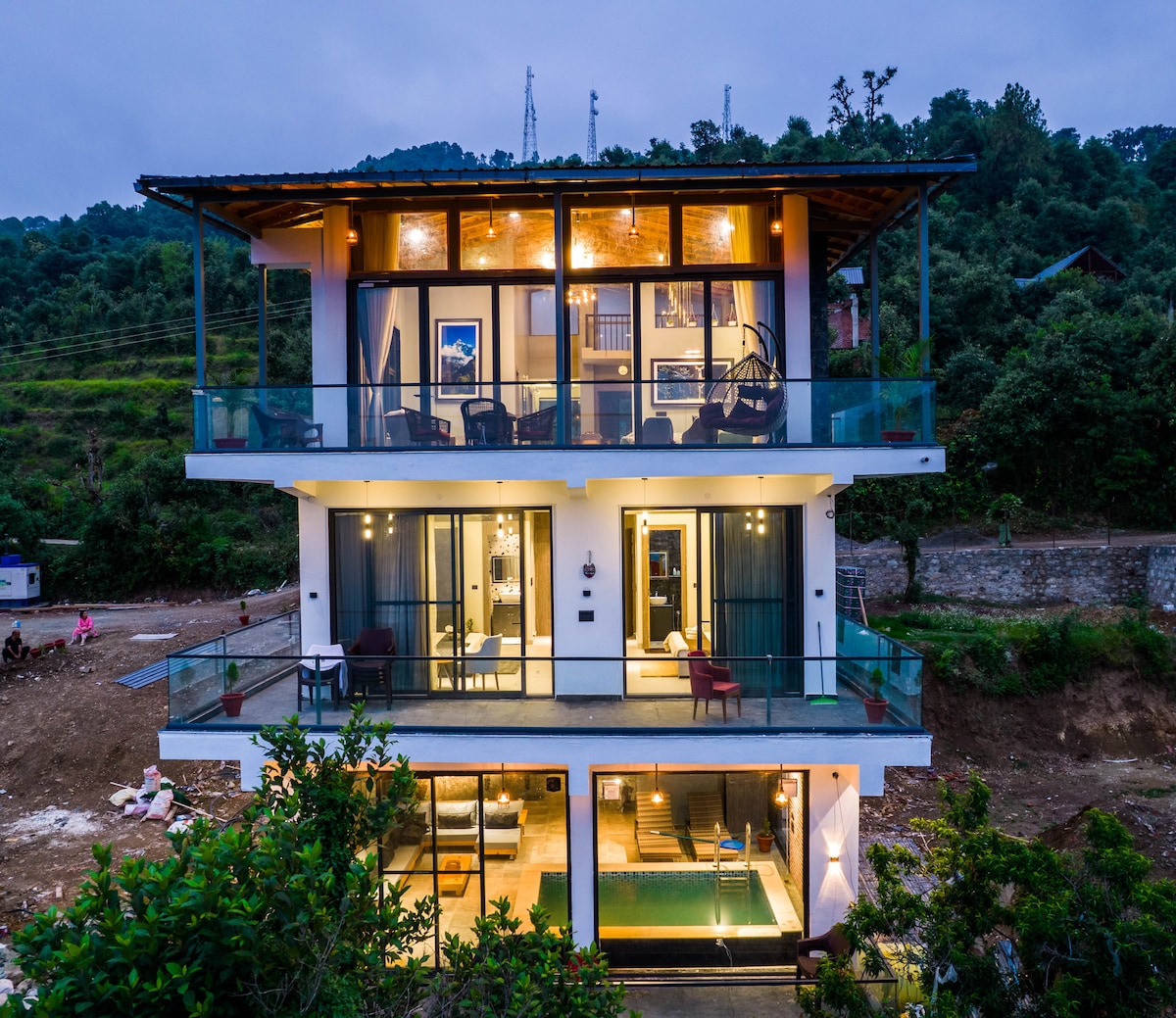 The three-story villa stands amidst a serene landscape, featuring large glass doors that provide panoramic views. Spacious balconies on each floor offer outdoor seating areas, and the warm glow of interior lights adds to the inviting appearance of the home.