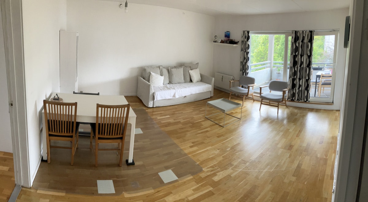 A spacious living area features a white sofa, light wooden dining table with four chairs, and a glass coffee table. Large windows allow natural light to fill the room, with a view of the green garden accessible from the balcony.