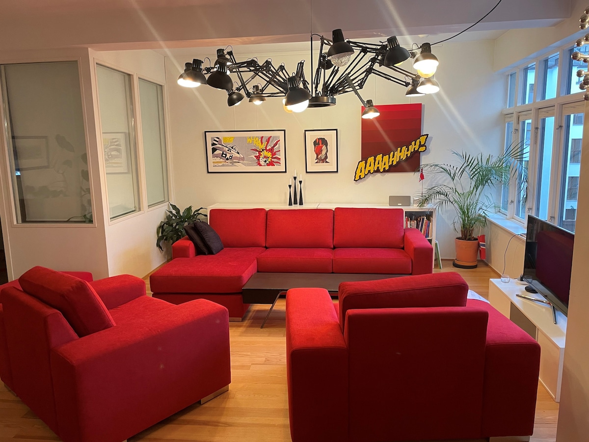 A spacious living area features a collection of red sofas arranged around a central coffee table. Bright artwork adorns the walls, complemented by varied lighting fixtures that provide ample illumination. Large windows invite natural light and are accompanied by indoor plants, enhancing the vibrant atmosphere.