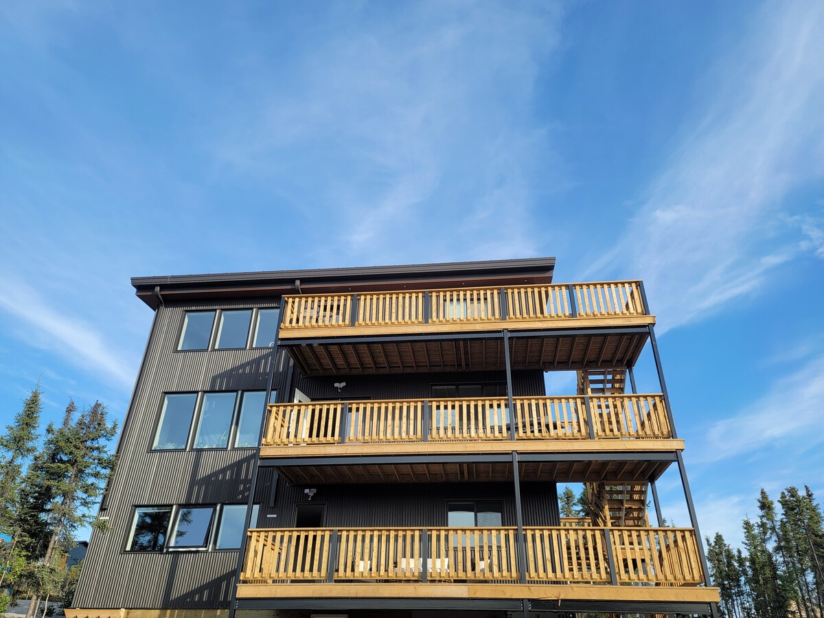 A three-story building is presented, featuring multiple balconies with wooden railings. Large windows are visible, allowing natural light to enter. The surroundings include trees and a clear blue sky, enhancing the building's modern architectural design.