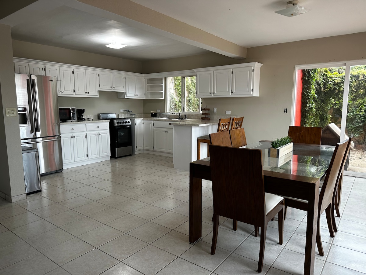 The bright kitchen features white cabinetry with modern appliances, including a stainless-steel refrigerator and gas stove. A large dining table with wooden chairs is positioned nearby, with natural light streaming through sliding glass doors that lead to an outdoor area.
