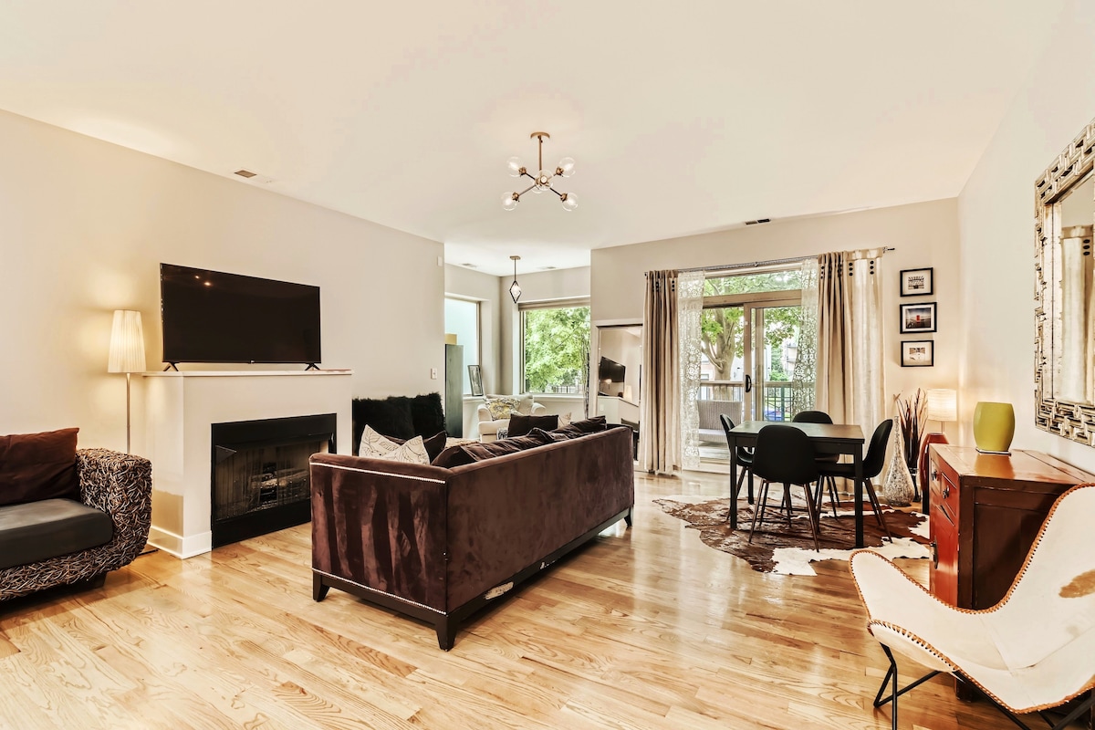 A spacious living area features a dark sectional sofa and a contemporary armchair. A flat-screen TV is mounted above a sleek fireplace. Natural light fills the space through large windows, which overlook greenery. A dining table with chairs is positioned nearby, accompanied by stylish decor.