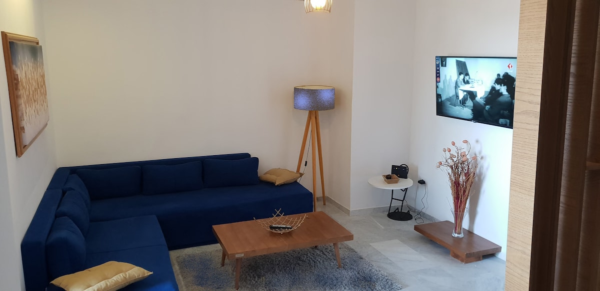 A living area features a blue sectional sofa adorned with gold cushions. A wooden coffee table sits in the center, complemented by a decorative bowl. A floor lamp provides soft lighting, and a wall-mounted TV is visible beside a small side table with a floral arrangement.