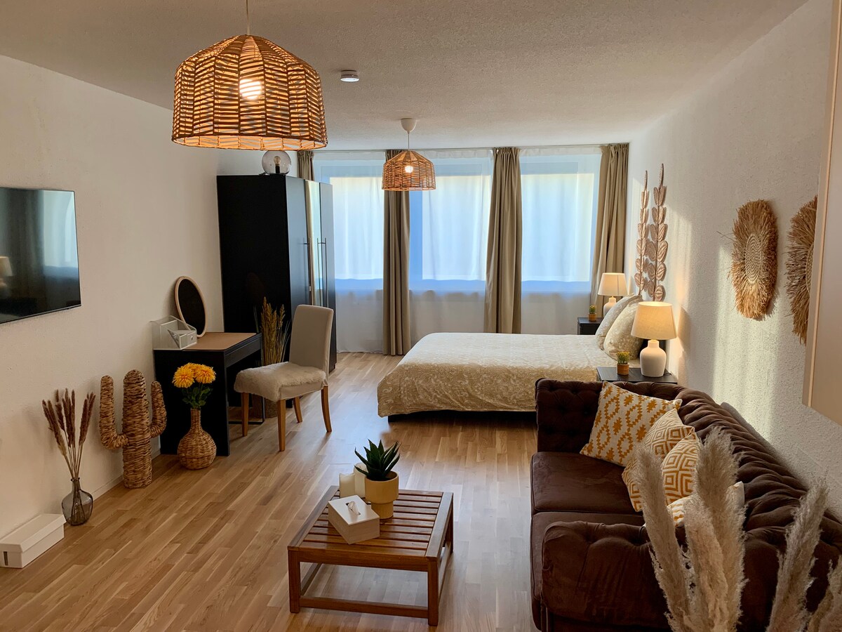 A modern studio space is presented, featuring a spacious bed adorned with soft bedding. Warm lighting from woven pendant lamps enhances the wooden floor. A comfortable sofa sits beside a low coffee table, while a small dining area includes a desk and chair. Large windows provide natural light.