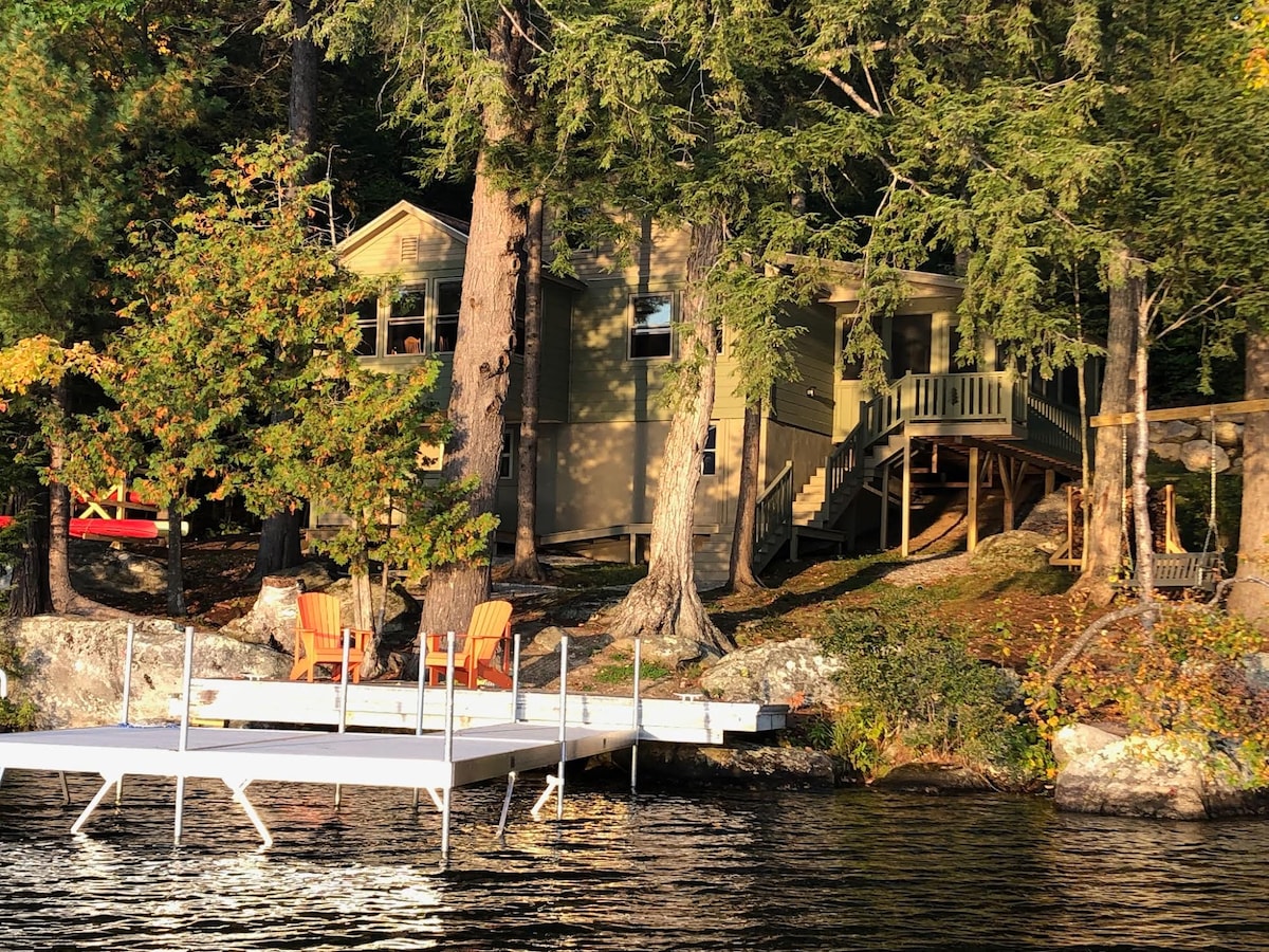 A lakefront house is set among tall trees, blending with the natural surroundings. A spacious dock extends over the calm water, featuring two Adirondack chairs. The entrance to the house is accessible via a set of steps leading to the deck.