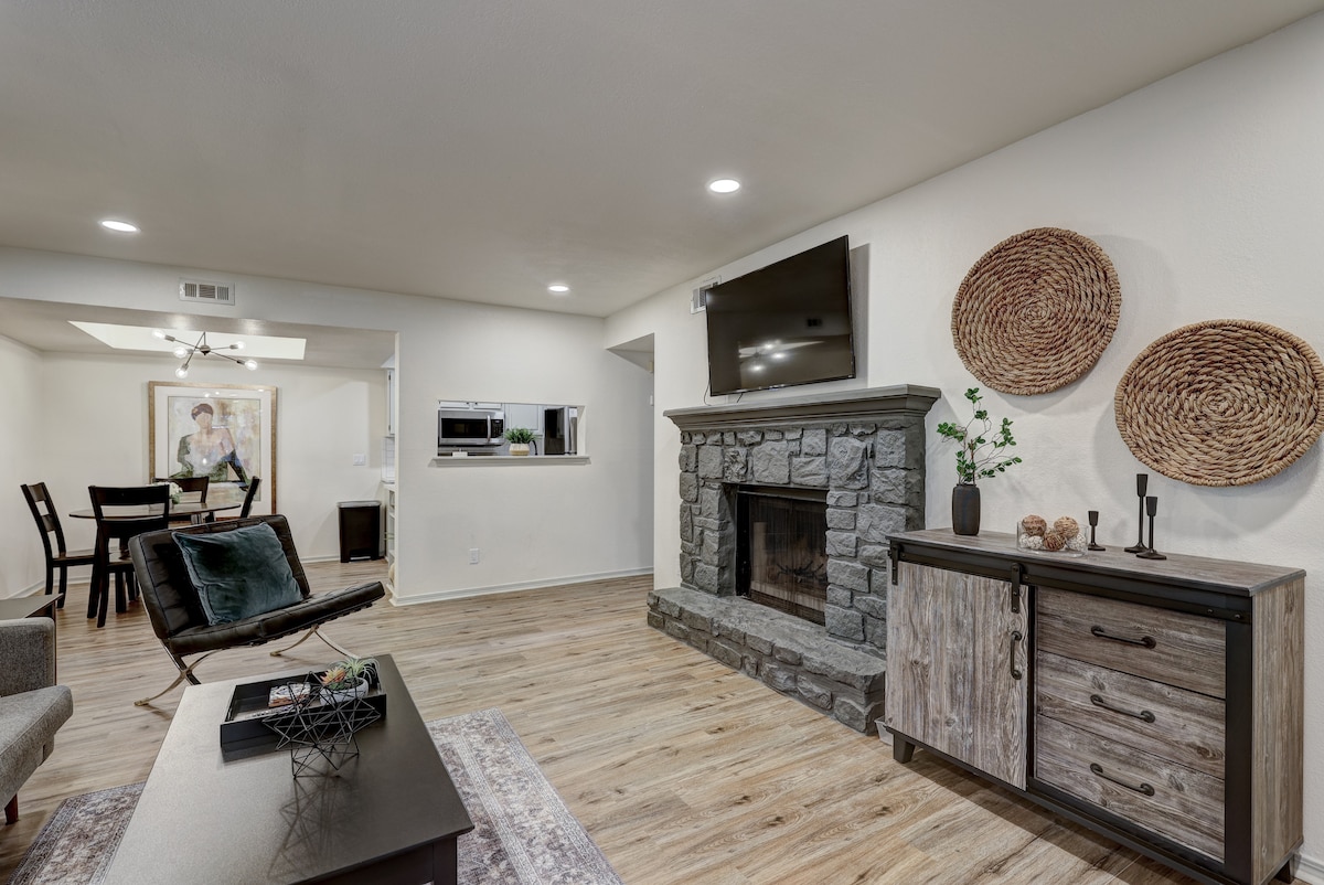 The living area features a stone fireplace as a focal point, accompanied by wooden storage and decorative elements. Natural light is provided by nearby windows, while a dining area with a black table and chairs is visible in the background, creating an open, connected space.