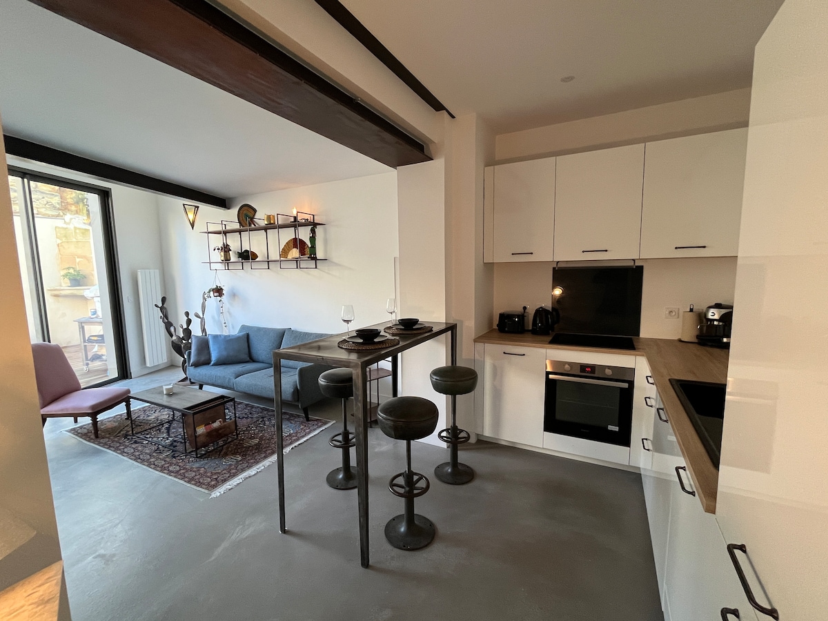 An open kitchen and living area features modern furnishings, including a blue sofa and tall black stools around a wooden dining table. Natural light fills the space through a large glass door, which leads to an outdoor area. Neutral tones are complemented by decorative items on the shelves.