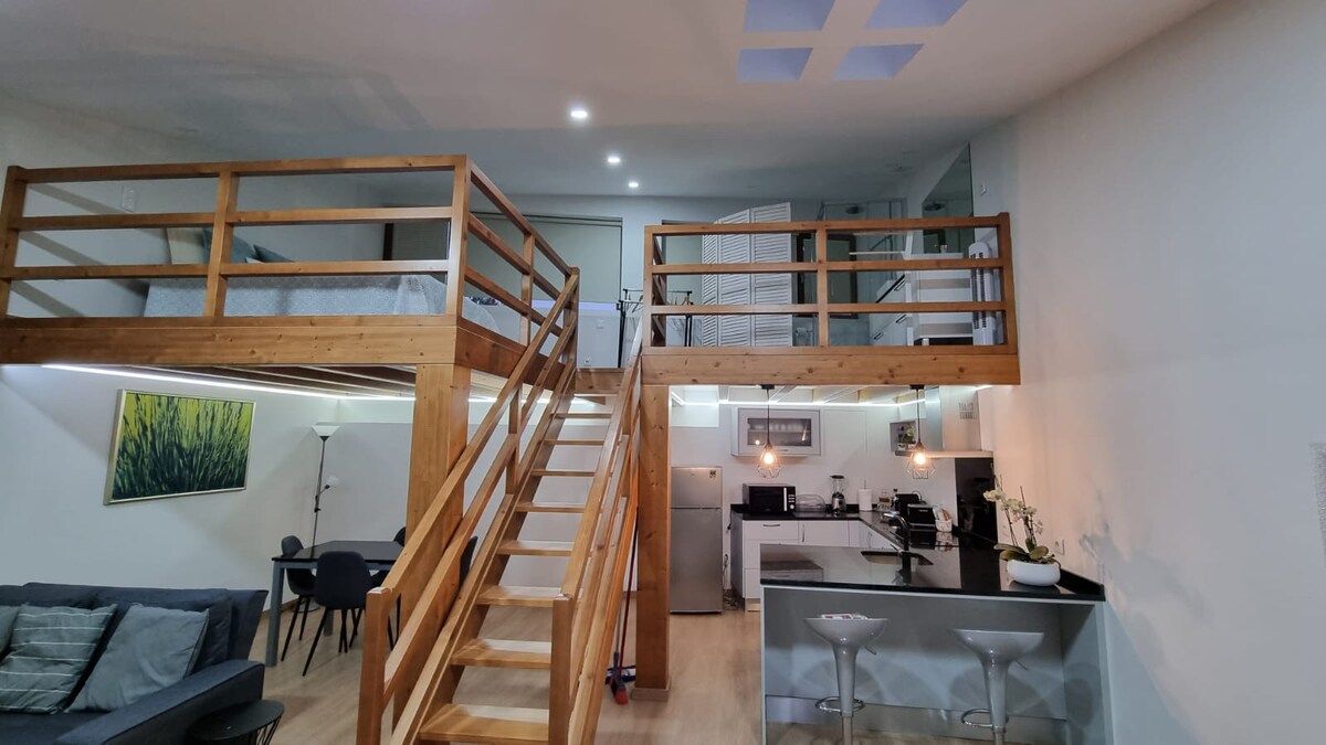 An open living space features a cozy seating area with a gray sofa and dining chairs. A wooden staircase leads to a lofted sleeping area, while the kitchen is equipped with modern appliances. Soft lighting enhances the overall ambiance of the space.