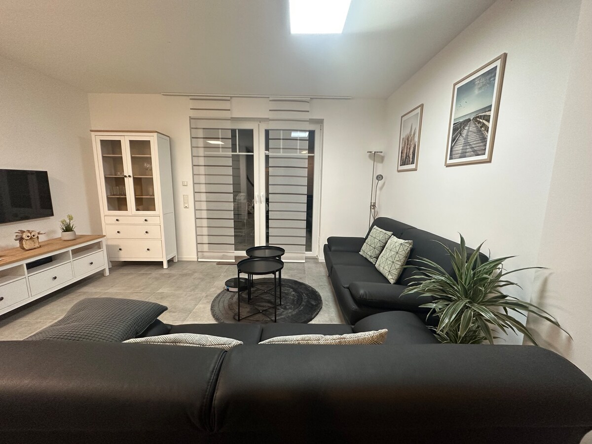 An open living area features a dark leather sofa and a round coffee table. Light-colored furnishings include a stylish cabinet and a flat-screen TV. Sliding glass doors allow natural light to enter, while decorative plants add a touch of nature to the space.