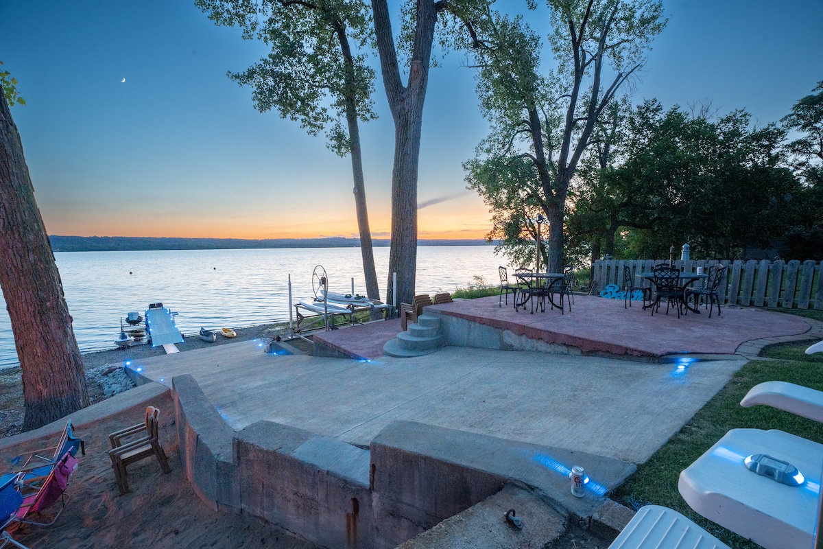 An expansive patio area is located by the lakeshore, featuring several tables and chairs arranged for outdoor dining. A tranquil view of the water and sunset is visible, with ambient lighting enhancing the setting. Large trees frame the scene, adding a natural touch.