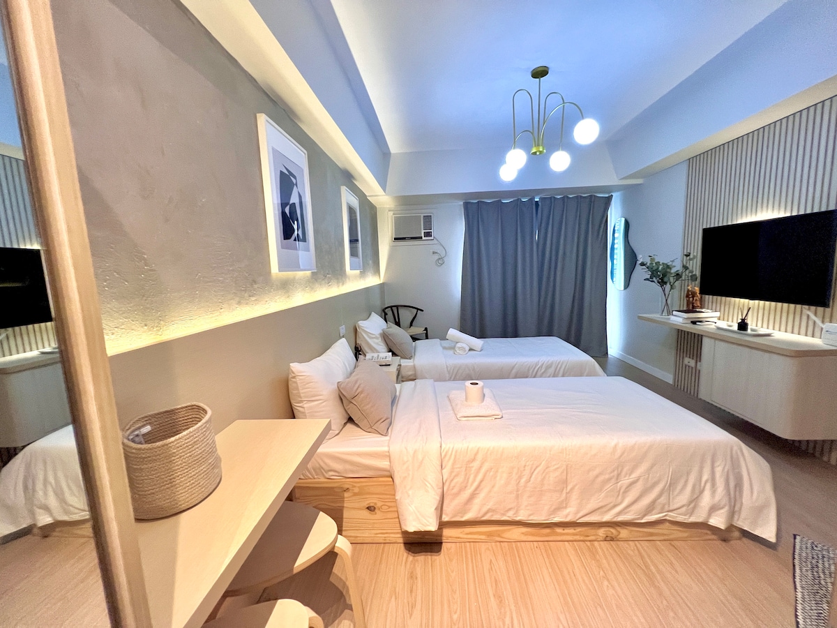 The studio unit features a bright, airy sleeping area with two single beds that can be converted into a king-size bed. A minimalist design is highlighted by wooden accents and modern decor. A wall-mounted TV and ample natural light enhance the welcoming atmosphere.