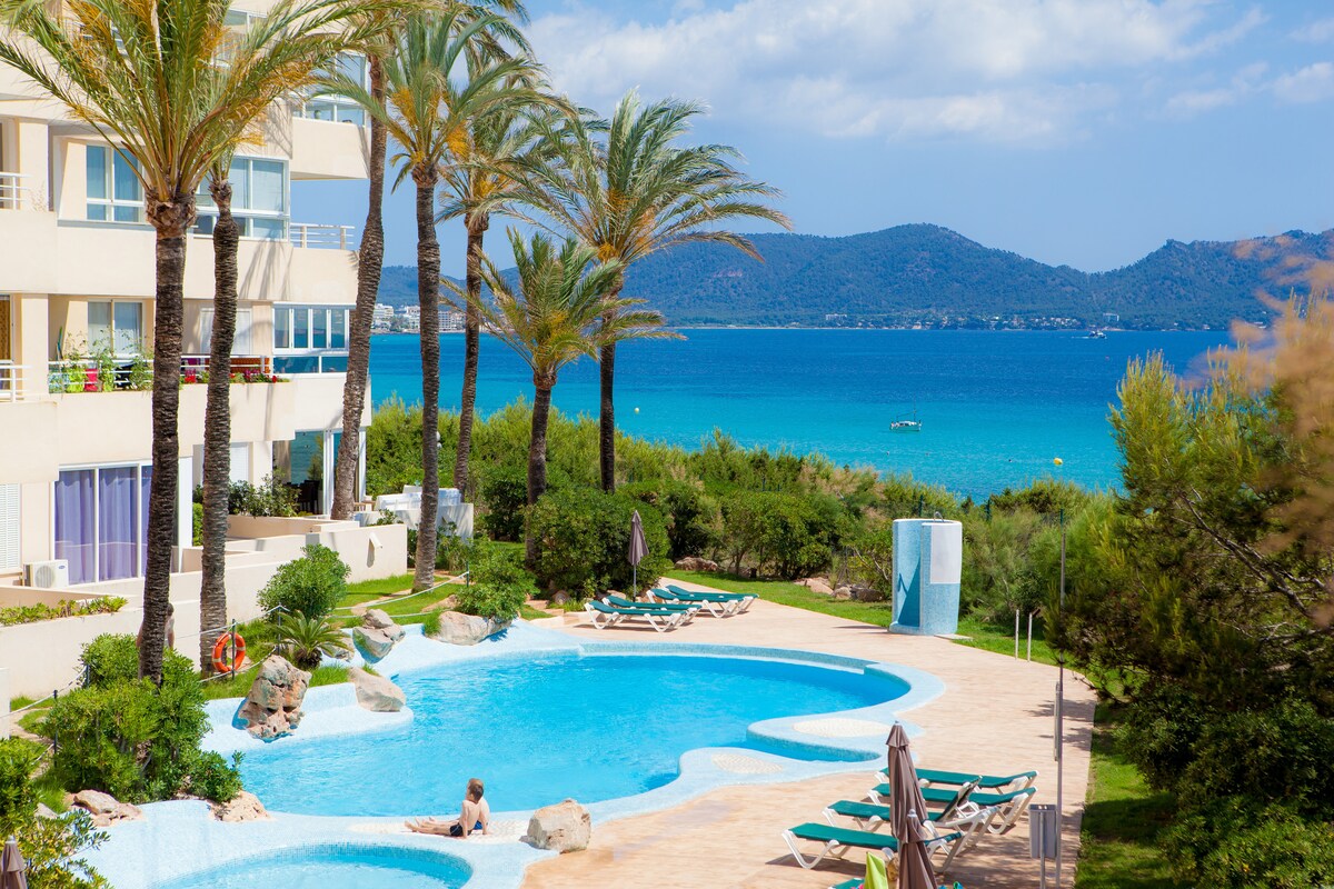Two inviting swimming pools are surrounded by lush greenery and palm trees, creating a relaxing atmosphere. The shimmering blue water reflects the sky. In the background, the beach and the sea are visible, offering a scenic view of the landscape.