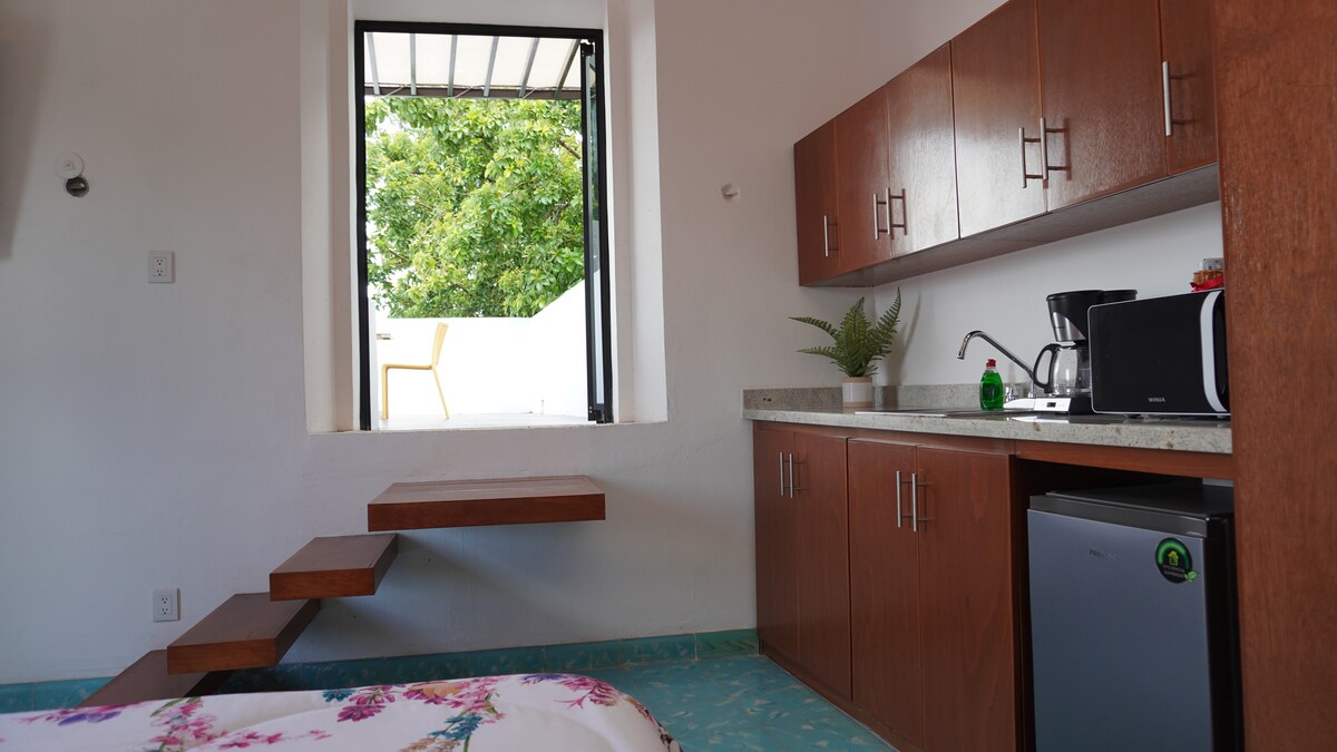 An inviting kitchenette is equipped with essential appliances, including a coffee maker and a refrigerator. A small wooden shelf extends from the wall, complementing the modern cabinets. Natural light enters through a window, framing a glimpse of the outdoor terrace.