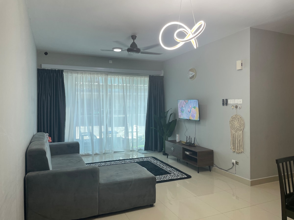 A modern living room features a gray sectional sofa positioned beside a sleek television stand. Large glass doors are dressed with sheer and dark curtains, allowing natural light to fill the space. Decorative elements include a minimalist wall clock and a plant, enhancing the ambiance.