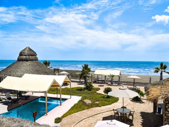 A view of the tranquil beach and ocean, framed by a thatched gazebo and sun loungers. The inviting pool area is surrounded by tropical greenery and features shaded cabanas. Clear skies and soft clouds enhance the serene coastal atmosphere.