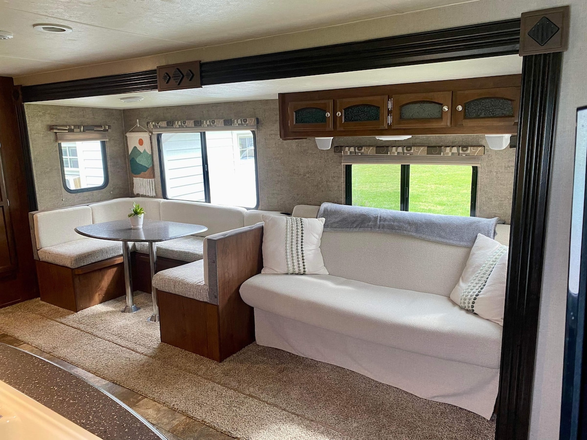 A comfortable seating area features a curved dinette with plush benches and a round table. A cozy sofa rests against the wall, complemented by natural light streaming through large windows lined with subtle curtains.