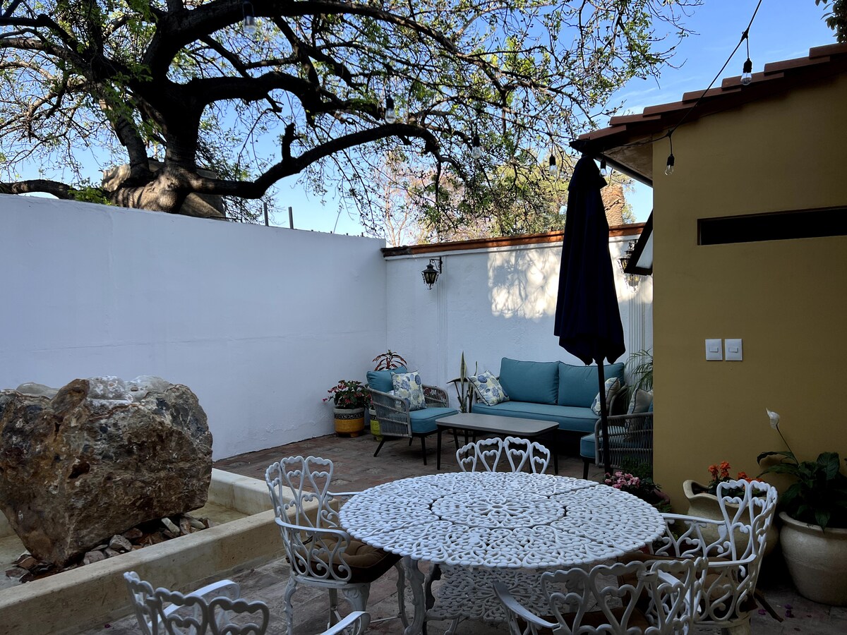 A spacious patio is showcased, featuring a decorative stone table surrounded by multiple white chairs. A comfortable seating area with blue cushions is positioned against one wall, and a large bougainvillea-covered tree provides shade. A small fountain is visible, enhancing the outdoor setting.