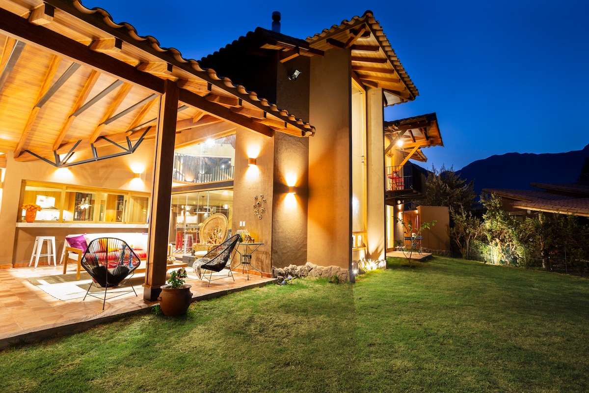 The exterior of Casa Bini is illuminated at night, showcasing a blend of rustic architecture and modern design. Outdoor seating areas are arranged on the lawn, with mountain silhouettes in the background, creating a serene ambiance.