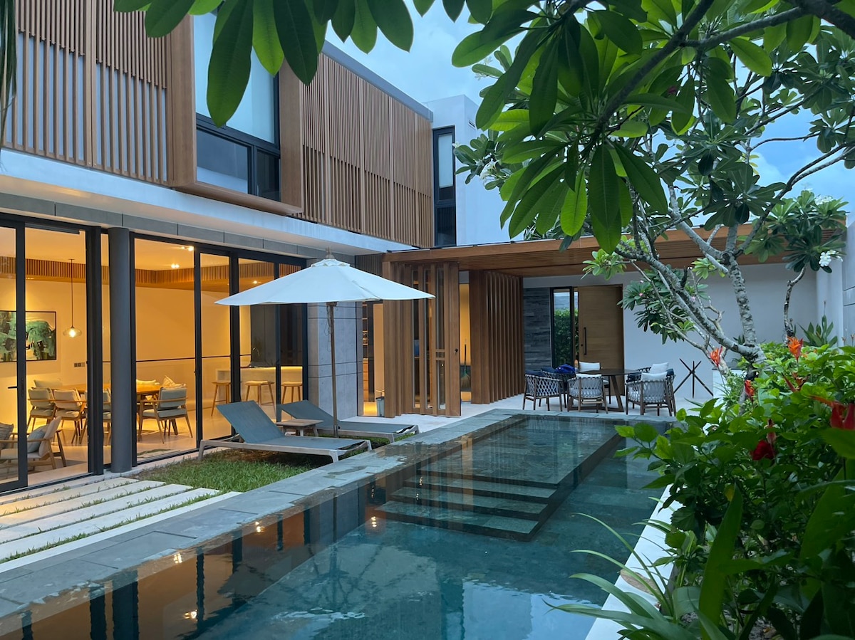 The modern villa is surrounded by lush greenery, featuring a private pool with steps leading into the water. A patio area is equipped with lounging furniture and an umbrella, while the interior space is visible through large glass doors, showcasing a dining area.