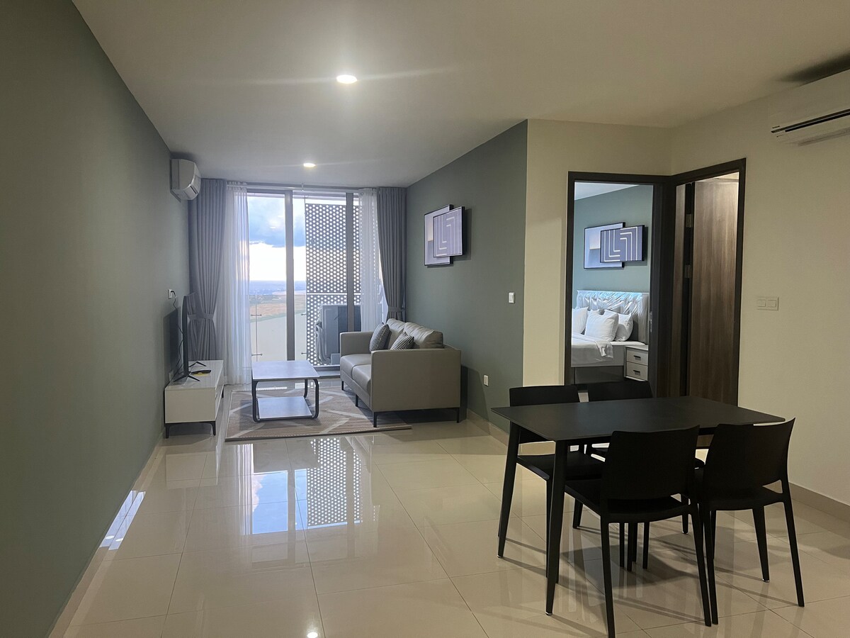 A spacious living area is visible, featuring a comfortable sofa and a television. A dining table with four chairs is positioned nearby. Natural light enters through large windows, illuminating the polished floor. A doorway leads to an adjoining bedroom area.