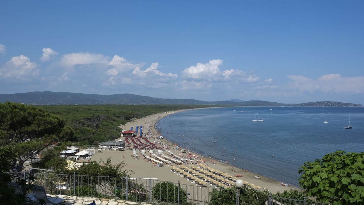 A long beach stretches along the shoreline, featuring a mix of areas with umbrellas and lounge chairs. The gentle slope into the water makes it accessible for families. A variety of people can be seen enjoying the beach under a clear blue sky, with scenic hills in the background.