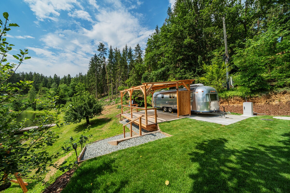 An original Airstream is positioned on a landscaped area featuring a wooden deck and gravel path. Lush greenery and trees surround the property, with the tranquil Haidensee visible in the distance. A shaded seating area complements the outdoor space.