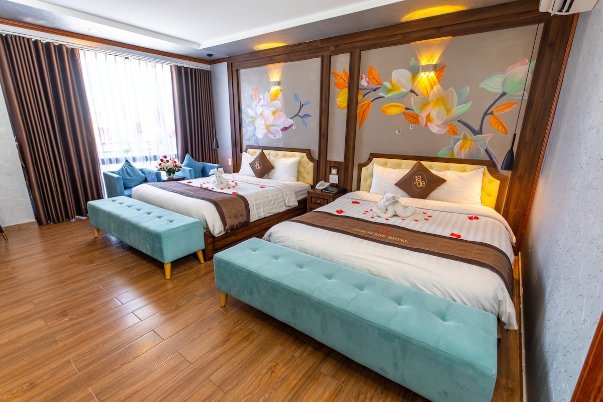 The room features two spacious beds with decorative pillows, placed against a mural adorned with floral designs. A soothing color palette of earth tones and soft blue accents is present. Large windows are dressed with dark curtains, allowing natural light to enhance the ambience.