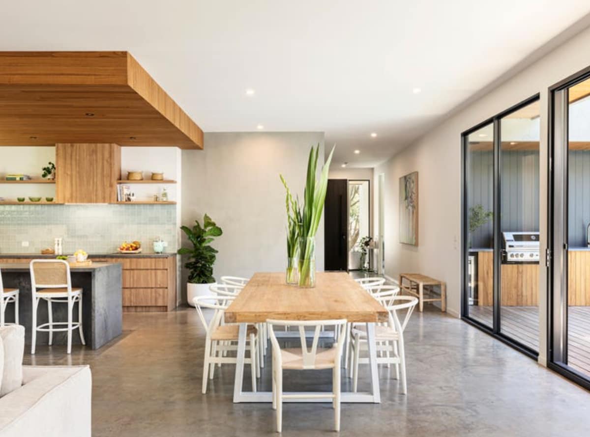 A spacious dining area features a large wooden table surrounded by white chairs. Natural light floods the room through expansive glass doors, with a view of the outdoor area, including a barbecue. A potted plant adds a touch of greenery to the neutral decor.