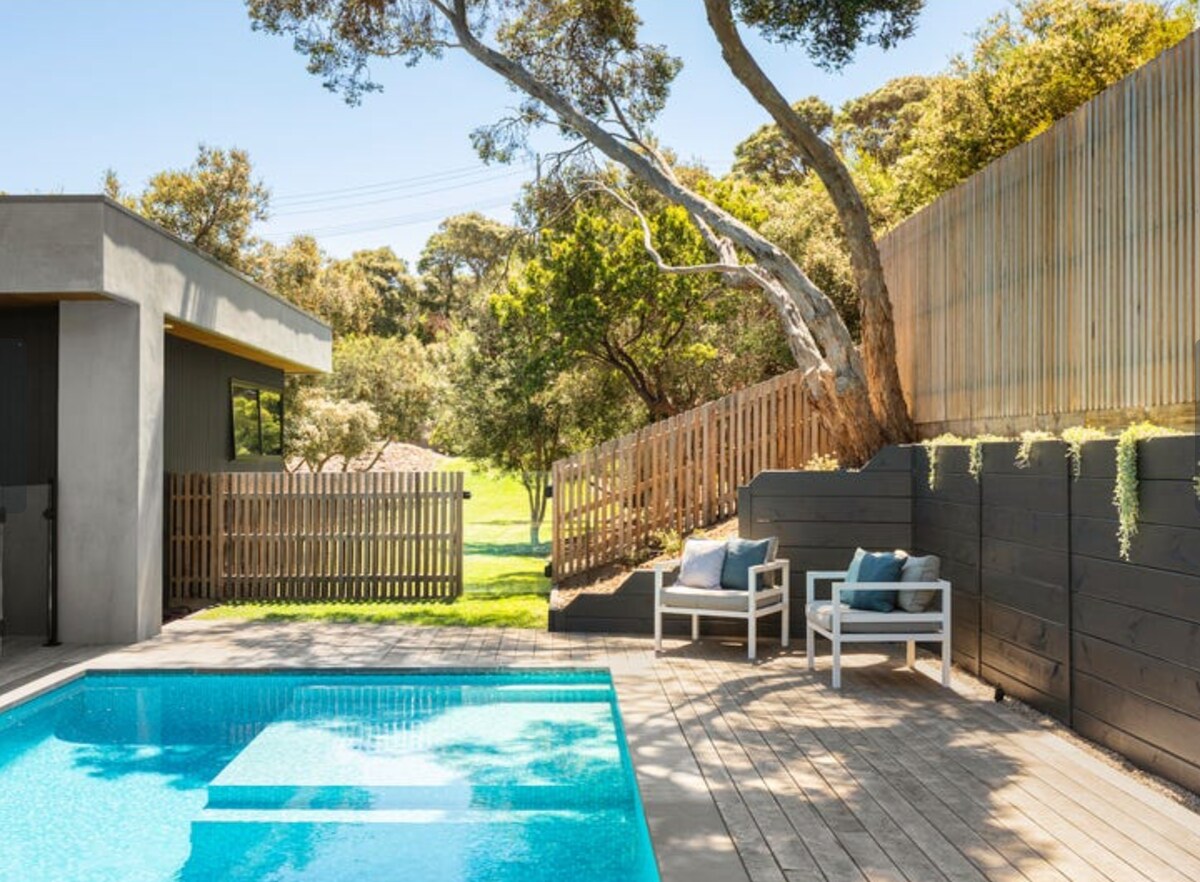 A shimmering solar-heated pool is framed by warm wooden decking and accented by minimalist white seating. Lush trees and greenery create a serene backdrop, with a wooden fence providing privacy. Bright sunlight casts a pleasant glow over the tranquil outdoor space.