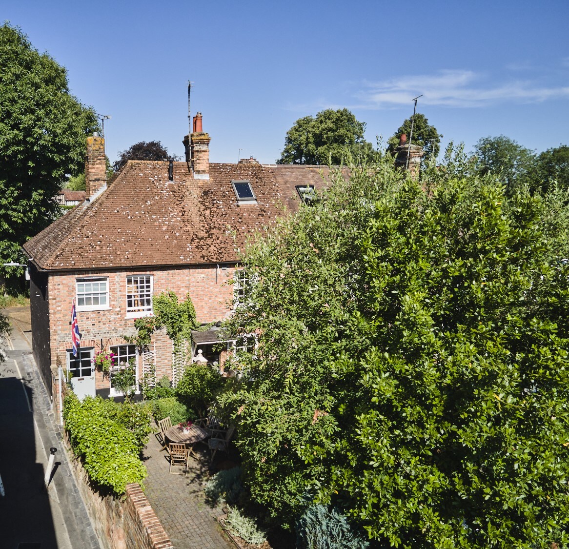The cottage is surrounded by lush greenery, featuring a charming brick exterior and a well-maintained garden. Outdoor seating is visible on the patio, providing a cozy spot to enjoy the natural surroundings. The clear blue sky complements the inviting atmosphere of this peaceful retreat.