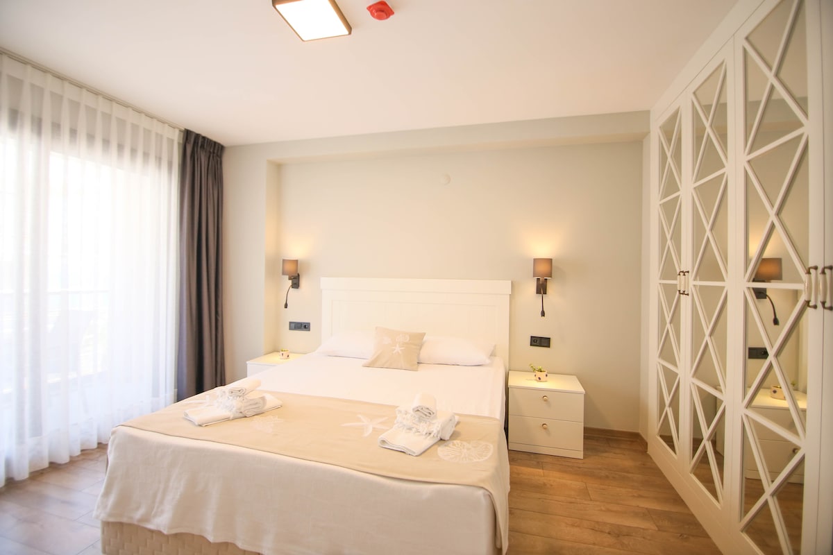 A light and airy bedroom features a spacious bed with an elegant cream blanket. Soft lighting is provided by wall sconces on either side. A large sliding wardrobe with a geometric design complements the room, while sheer curtains allow natural light to fill the space.