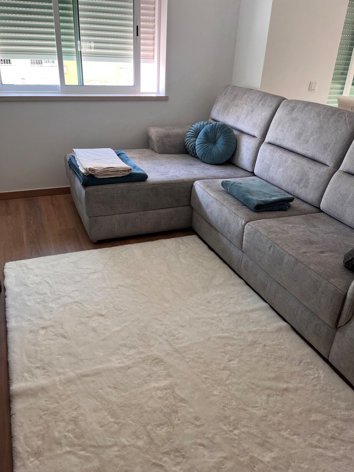 A light grey sectional sofa is positioned in a well-lit corner of the room, complemented by a textured white area rug. Soft, folded towels and a round cushion in teal are arranged on the chaise section, with sunlight filtering through the partially open window.