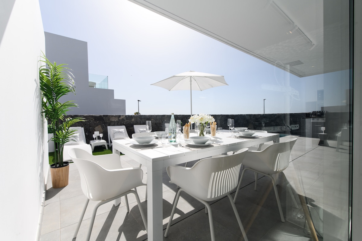 A dining area is displayed with a white table set for several guests, featuring dishes and glassware. An umbrella provides shade over the table. White chairs surround the table, and a potted plant adds a touch of greenery. Large glass doors offer access to the outdoors.