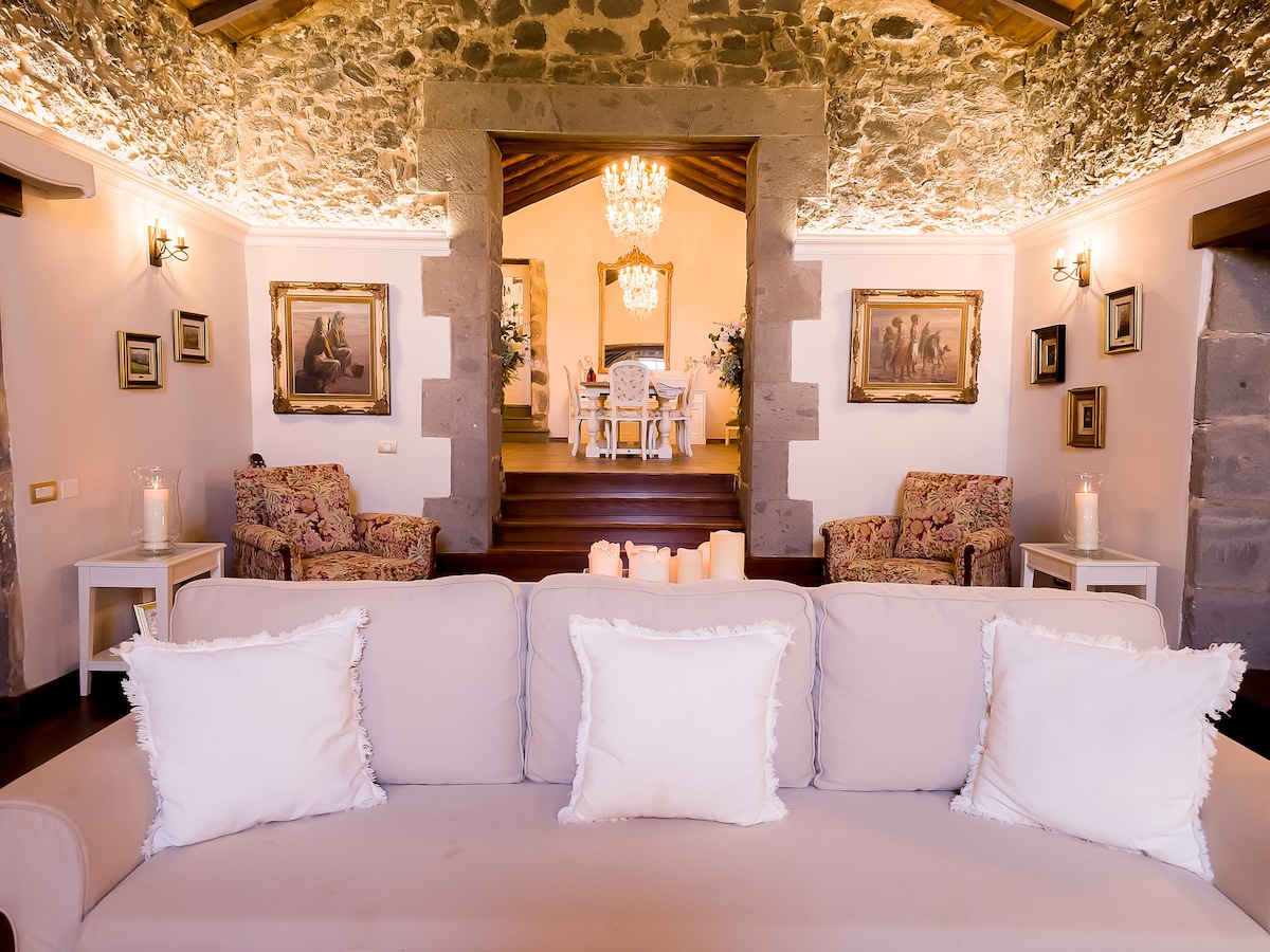 A cozy living area features a light-colored sofa adorned with decorative pillows, positioned in front of a stone archway. Elegant chandeliers illuminate the space, highlighting artwork on the walls and leading toward a dining area visible in the background.