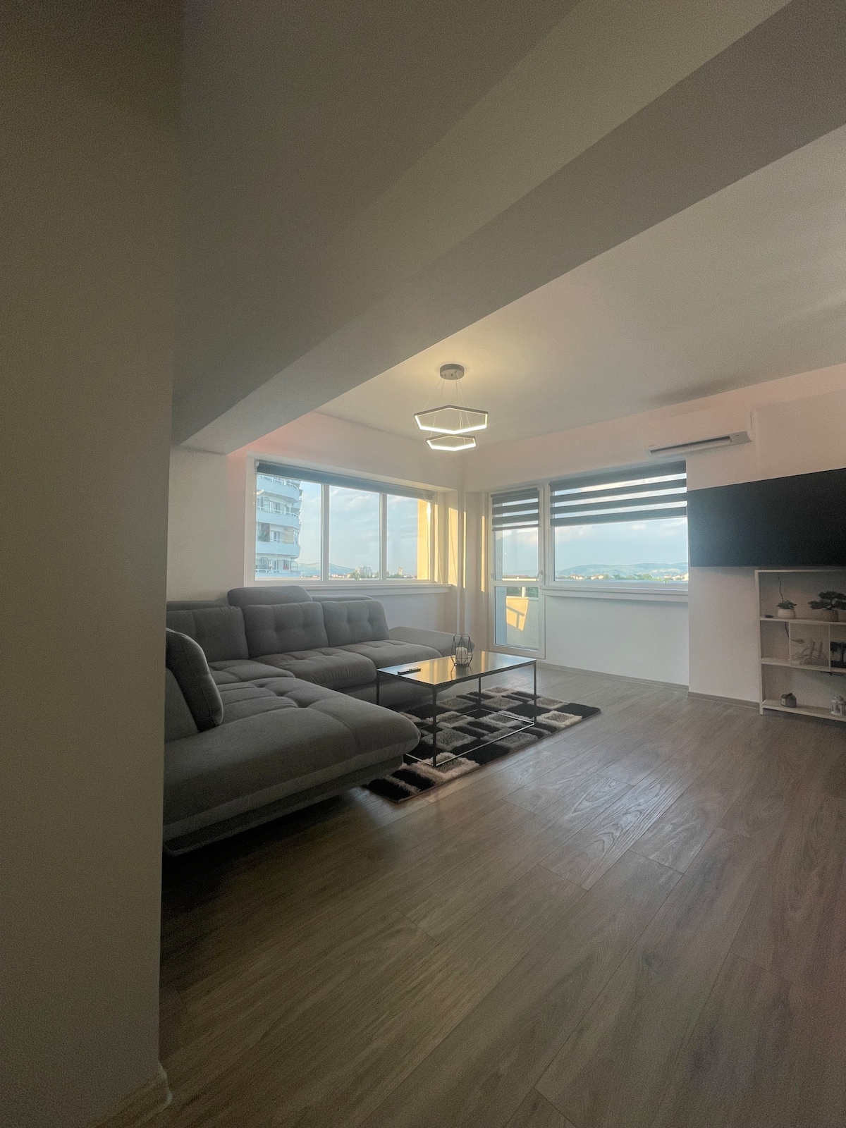 A modern living space is highlighted by large windows that allow natural light to fill the room. A gray sectional sofa is positioned beside a sleek glass coffee table, with a television mounted on the wall. Light wood flooring enhances the open and airy feel.