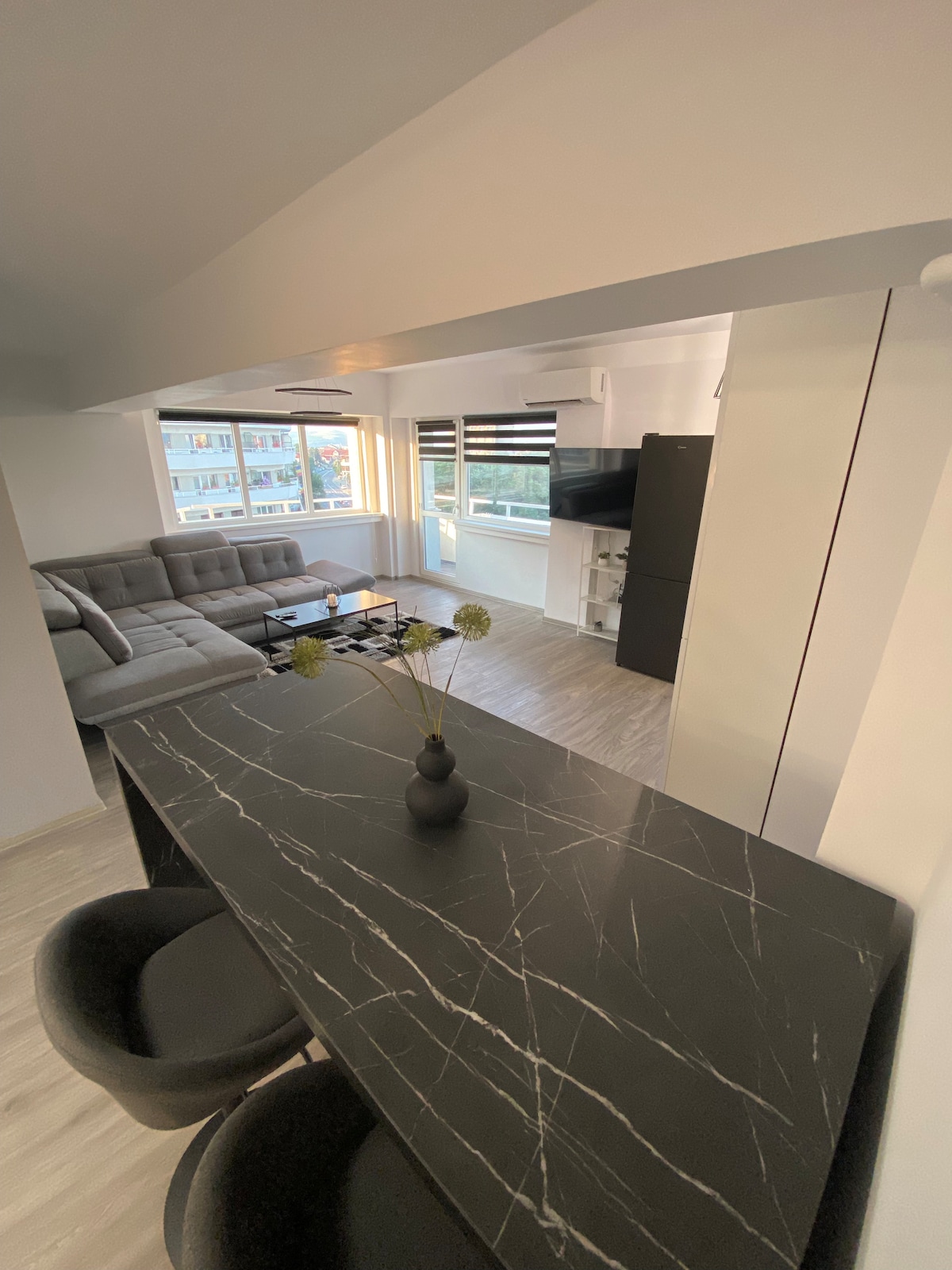 A modern living space is highlighted by a sleek dark marble table with two black chairs at the forefront. In the background, a spacious gray sofa faces a large window, allowing natural light to fill the room. Minimalist decor adorns the walls.