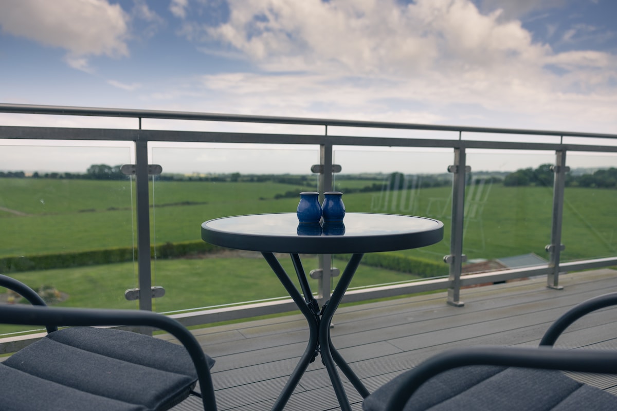 A balcony with a round table and two chairs overlooks expansive green fields. Two blue vases are placed on the table, complementing the natural surroundings. The railing offers unobstructed views of the landscape, providing a serene outdoor seating area.