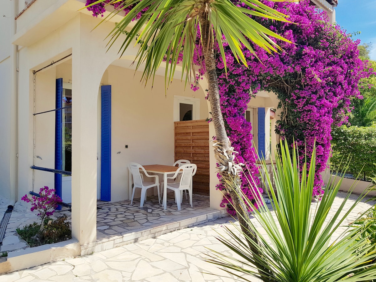 A private terrace features a wooden table surrounded by white plastic chairs, with vibrant bougainvillea creating a natural overhead canopy. The sunlit space is bordered by a stone pathway and lush tropical plants, offering a relaxing outdoor setting.