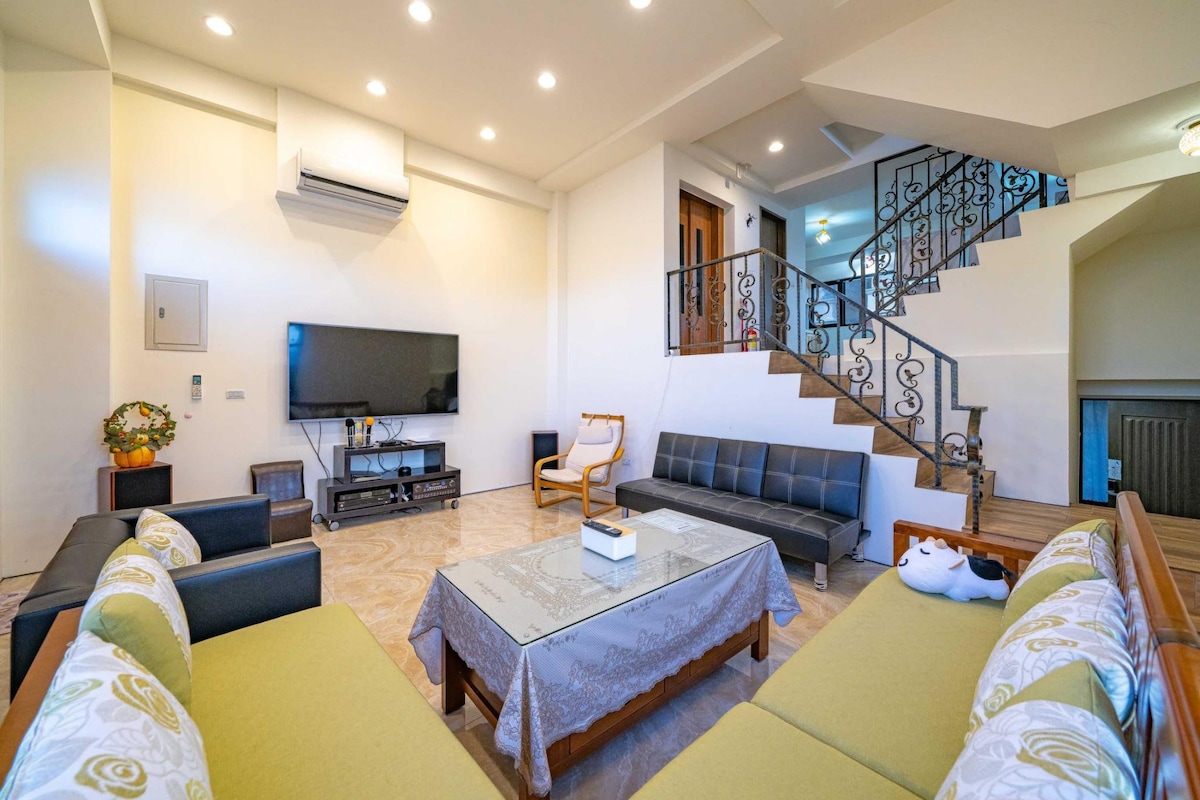 A spacious living area features a large flat-screen TV mounted on the wall, with comfortable seating arrangements that include a plush sofa and armchairs. A decorative coffee table occupies the center, and natural light floods in from the windows, enhancing the welcoming ambiance.