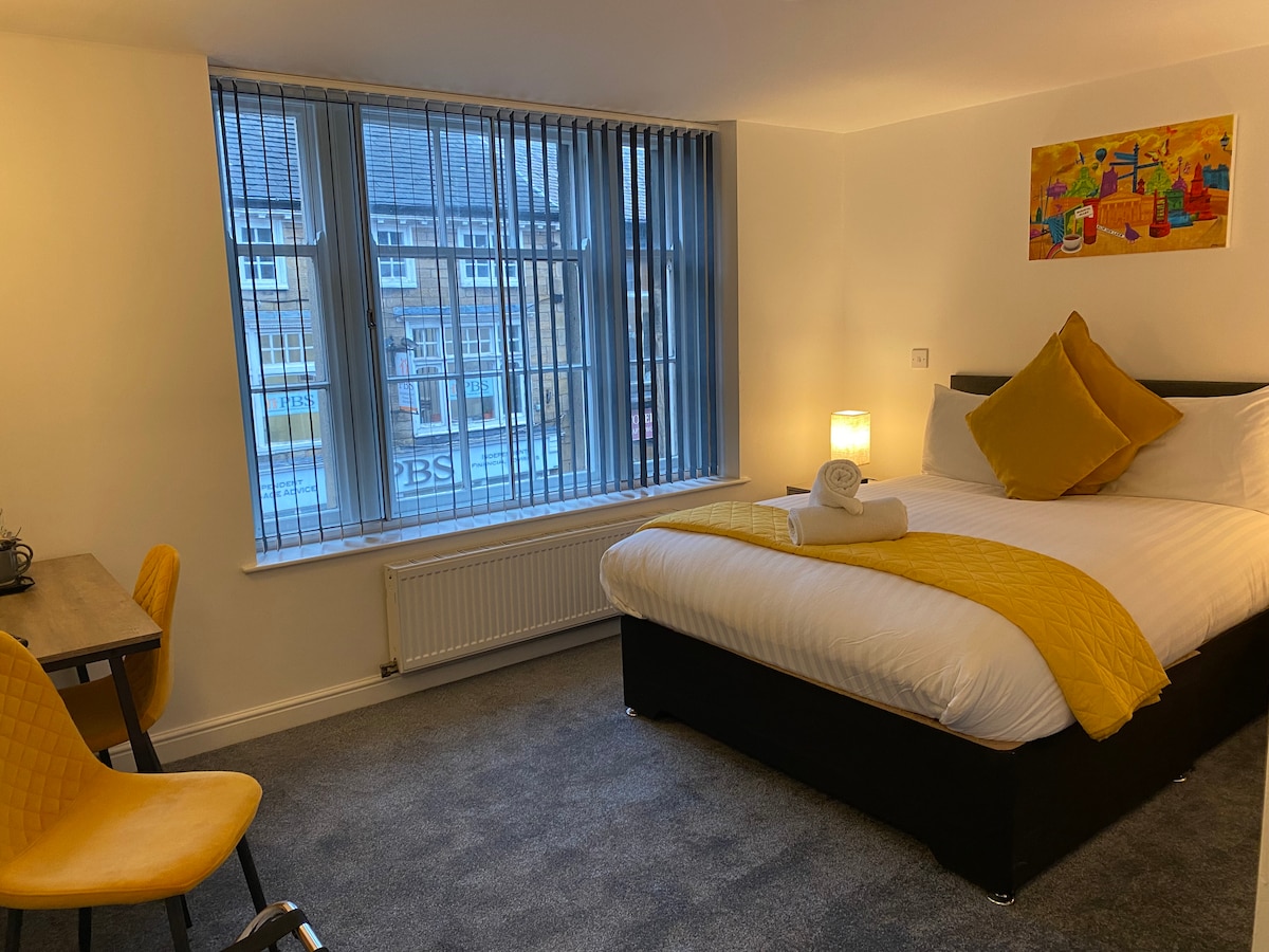 A well-appointed double room features a spacious bed with a bright yellow throw and decorative cushion. A small desk with an orange chair is positioned beside a large window, allowing natural light to fill the room. Soft carpeting and simple wall art complete the inviting ambiance.