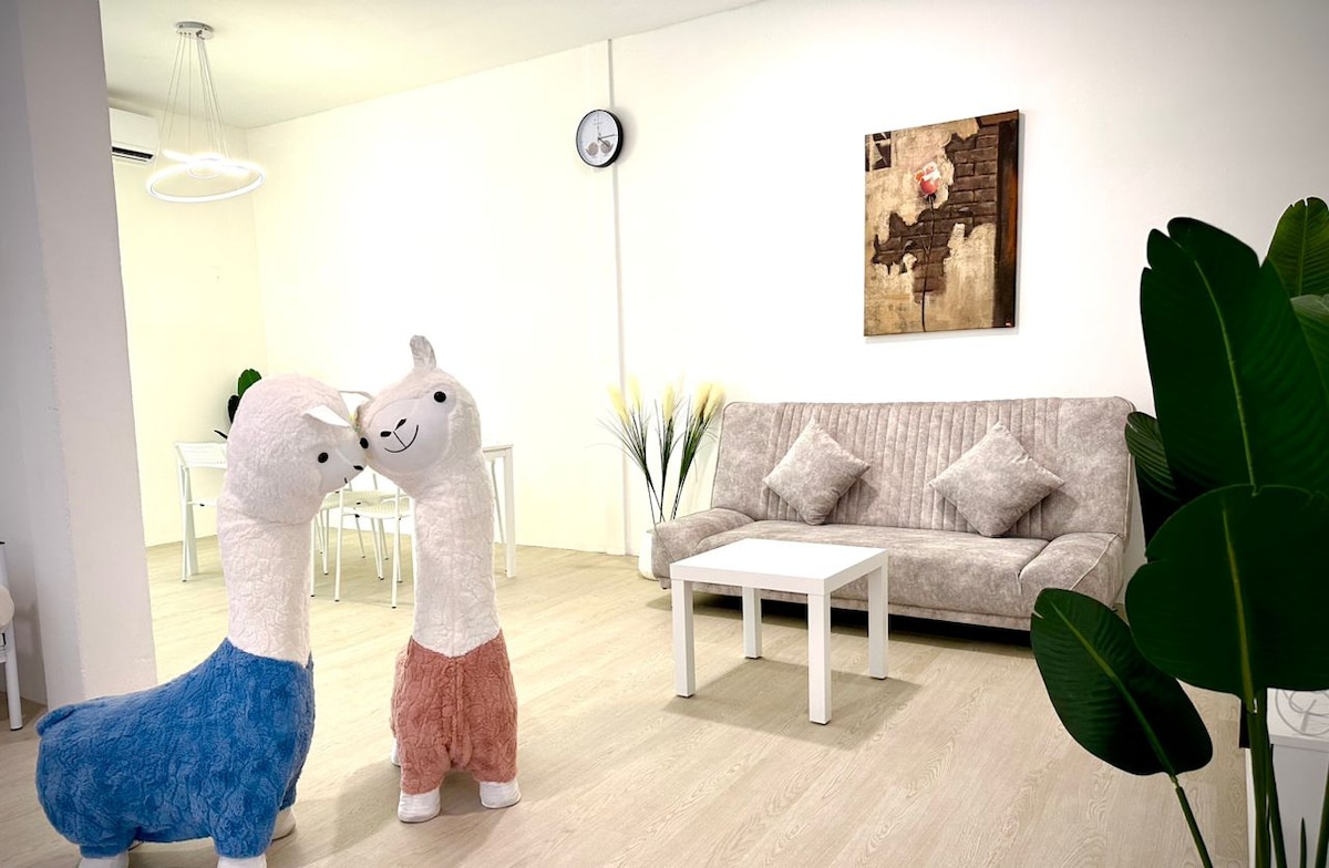 Two plush llama decorations stand playfully in a bright, minimalistic living area. A textured sofa is positioned against the wall, accompanied by a simple coffee table. A wall clock and a piece of art provide subtle accents, while soft greenery adds a touch of nature.