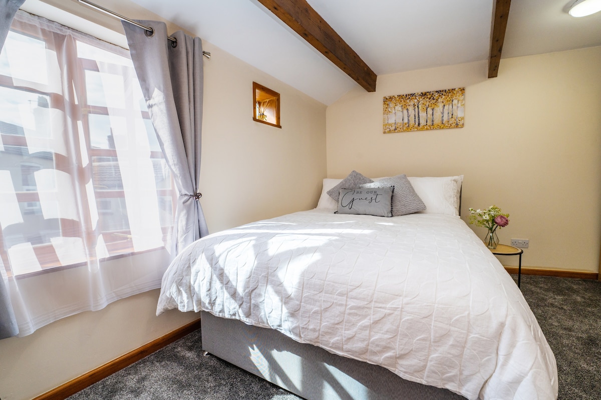 A well-lit bedroom features a spacious bed with a white quilt and decorative pillows. Natural light filters through a window draped with sheer curtains. Exposed wooden beams add character, while a framed piece of art and a vase with flowers are displayed on the wall.